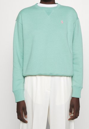 Personne portant un sweat-shirt vert menthe à manches longues avec un logo rose brodé et un pantalon blanc taille haute sur un fond uni.