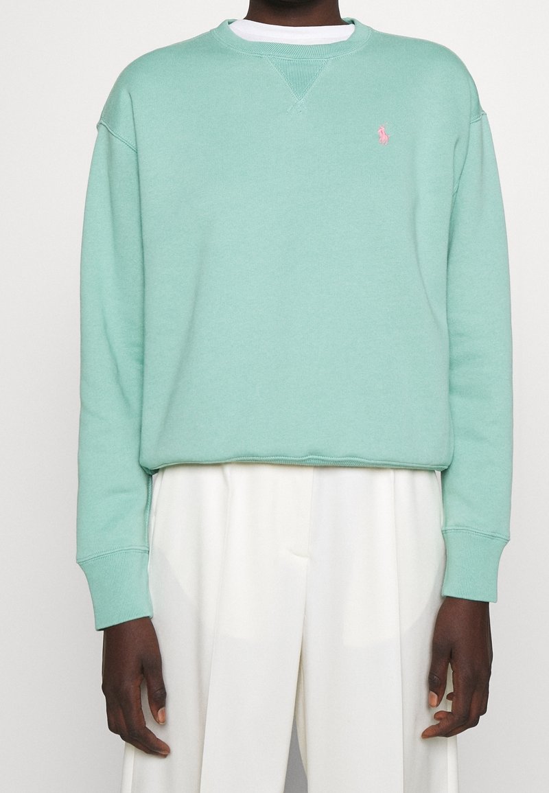 Personne portant un sweat-shirt vert menthe à manches longues avec un logo rose brodé et un pantalon blanc taille haute sur un fond uni.
