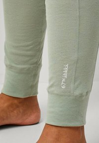 Untere Beine in hellgrünen Jogginghosen mit gerippten Bündchen und weißem "67°N Áhkká" Branding in der Nähe des rechten Knöchels, barfuß auf einem grauen Boden.