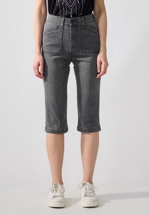 Person iført grå denimshorts i midtlængde med store forlommer og hvide sneakers, stående mod en ensfarvet lys baggrund.