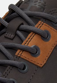 Close-up of dark gray shoe with thick gray laces threaded through black metal eyelets and brown leather patch detail.