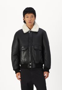 Černá kožená bomber bunda s bílým ovčím límcem, dvěma předními kapsami, žebrovanými manžetami a knoflíkovým zapínáním. Hladká textura po celém obvodu.