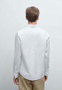 Une personne jeune avec les cheveux bruns courts se tient face à un mur gris clair uni, portant une chemise blanche à manches longues et un pantalon beige.