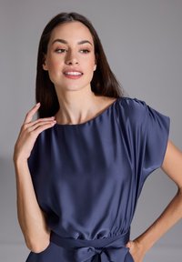 Robe en satin bleu marine avec un décolleté bateau, des manches courtes et un détail de taille nouée. La texture lisse et la brillance subtile mettent en valeur le design.