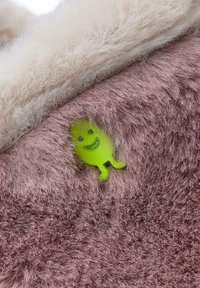 Un tissu rose flou avec un doux bord blanc présente un petit personnage en plastique vert souriant, intégré dans le matériau.