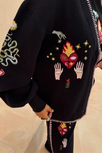Čierny pletený kardigán s farebnými vyšívanými dizajnmi, vrátane rúk, srdcí, hviezd a hadov. Obsahuje kontrastný zips a prešívanie.
