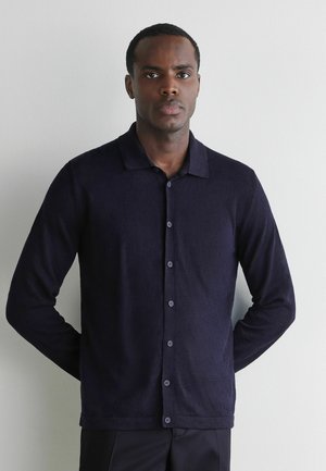 Mann mit kurzen Haaren, der ein dunkelblaues, langärmeliges Hemd mit Knöpfen trägt, steht vor einer schlichten hellen Wand, die Hände hinter dem Rücken.