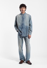 Jasnoniebieska koszula dżinsowa i spodnie w luźnym kroju. Koszula ma zapięcie na guziki i klasyczny kołnierzyk. Całość dopełniają brązowe buty.