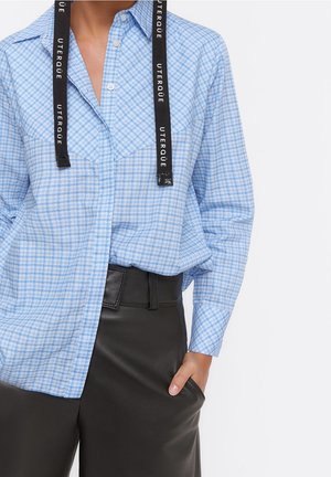 Blaue karierte Hemdbluse mit schwarzen Logo-Trägern, langen Ärmeln und lockerer Passform, kombiniert mit schwarzen hochtaillierten Hosen.