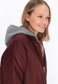 Chaqueta bomber color burdeos con capucha gris. Cuenta con un cierre frontal con cremallera y puños acanalados. Tejido liso y brillante con un diseño informal.