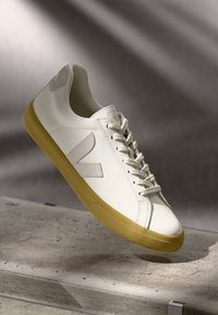 Hvit lær sneakers med gul gummisåle, tonede lisser og grå detaljer på hælen; har et merket logo og minimalisk design.