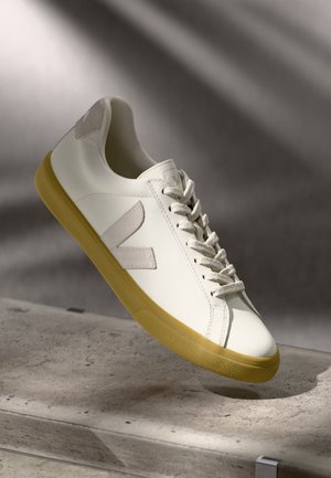 Sneaker in pelle bianca con suola in gomma gialla, lacci tono su tono e dettagli grigi sul tallone; presenta un logo di marca e un design minimalista.