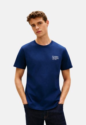 Joven con una camiseta azul marino Tommy Hilfiger con el logo en el pecho izquierdo, manos en los bolsillos, sobre un fondo claro y liso.