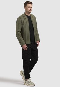 Oliven grøn quiltet jakke med lige snit, lange ærmer og lynlås foran, parret med sorte cargo bukser og lyse sneakers.