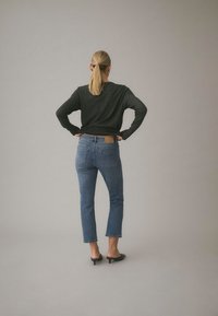 Femme avec une queue de cheval blonde portant un t-shirt noir à manches longues, un jean bleu et des mules noires à talons, debout les mains sur les hanches, face à un fond uni.