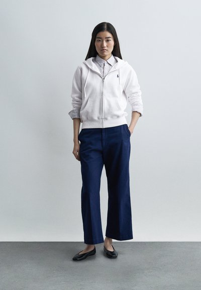 Giovane donna in piedi con le mani nelle tasche di pantaloni larghi blu navy, indossa una felpa bianca con zip sopra una camicia a righe con colletto e scarpe basse nere.