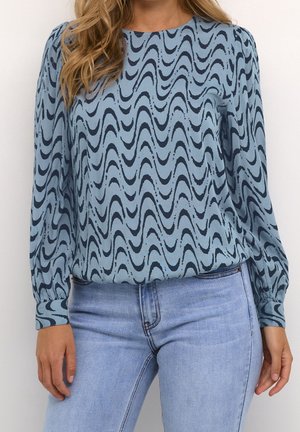 Femme portant un chemisier bleu à motif de lignes ondulées noires et un jean bleu clair sur un fond uni.