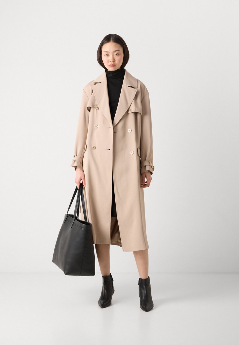 Beige Trenchcoat mit doppelreihigen Knöpfen, breiten Revers und Manschetten-Details. Kombiniert mit einem schwarzen Oberteil, einer schwarzen Tote-Tasche und schwarzen Knöchelstiefeln.