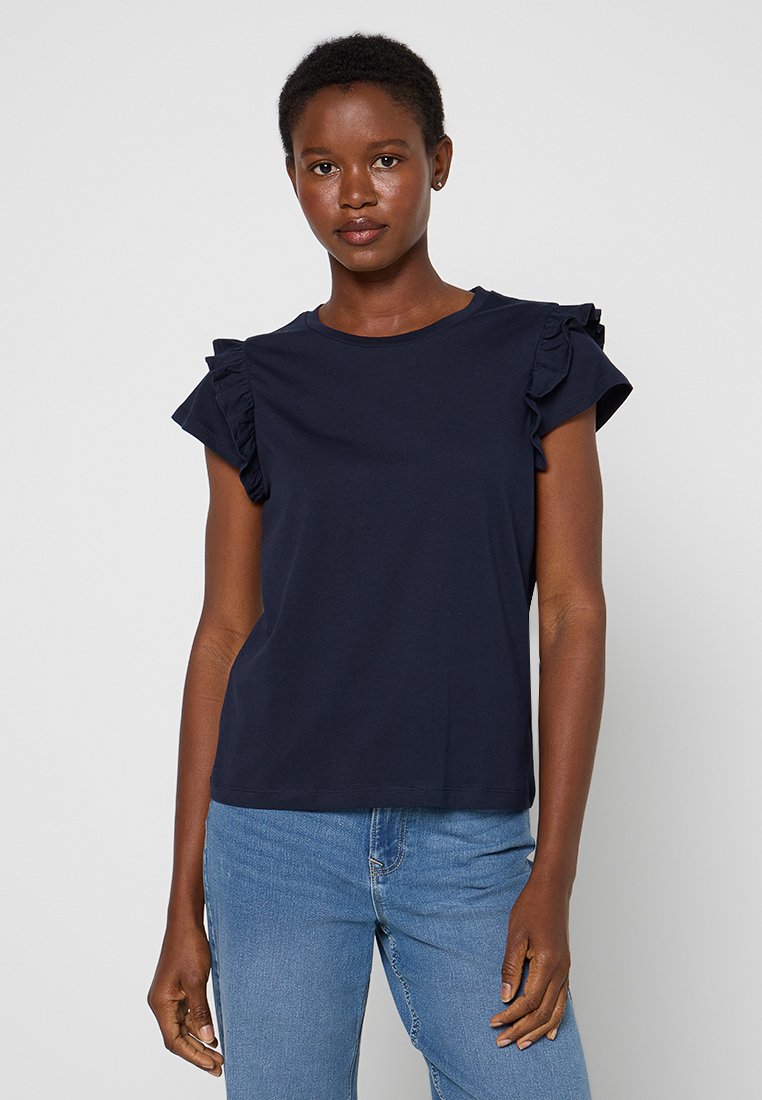 Frau mit kurzen Haaren, die ein navyblaues Oberteil mit Rüschensäumen und hellblaue Jeans trägt und vor einem schlichten hellen Hintergrund steht.