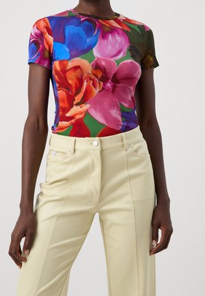 Osoba ubrana w żywą, kwiecistą bluzkę z krótkim rękawem z dużymi różowymi, niebieskimi i pomarańczowymi kwiatami, w połączeniu z wysokim stanem beżowych spodni.