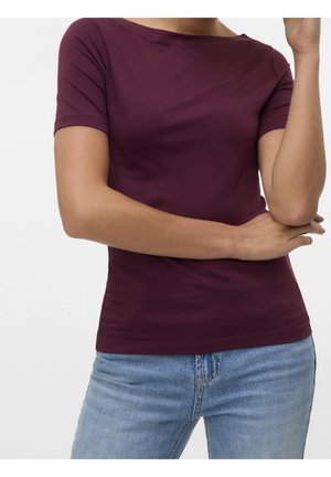 Vrouw met een aansluitend bordeauxrood topje met korte mouwen en lichtblauwe jeans, met één arm gekruist en de andere omhoog gebogen.