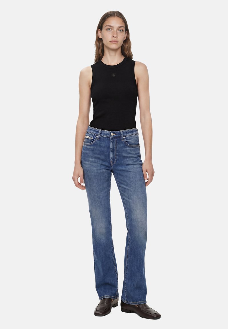Femme debout portant un haut noir sans manches, un jean bleu en denim et des chaussures marron foncé à bout carré, sur un fond blanc uni.