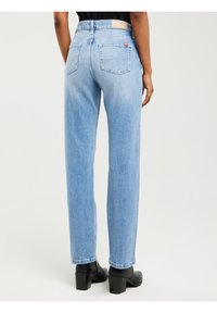 Naine, kes kannab helesiniseid sirge säärega teksapükse ja musti kontsaga pahkluu-saabasteid, seisab ühevärvilise hele tausta ees.