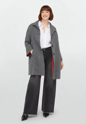 Manteau oversize tissé gris et noir avec un col haut, des poches latérales et une doublure rouge, associé à un jean noir à jambes larges et des talons noirs.