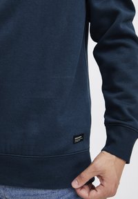 Donkerblauwe sweatshirt van een zachte, gestructureerde stof met geribbelde mouw- en onderkanten. Heeft een klein zwart label nabij de taille.