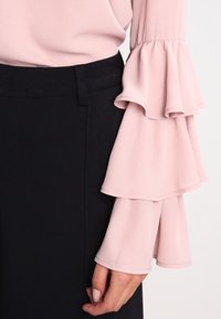 Blusa rosa con maniche a volant, realizzata in tessuto leggero. Indossata con una gonna nera, che mostra una texture liscia e un design a strati.