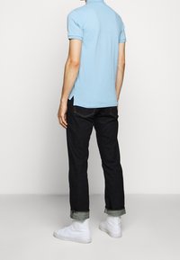 Man som har på sig en ljusblå kortärmad polo, mörka uppvikta jeans och vita högskaftade sneakers och står mot en enfärgad bakgrund.