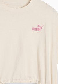 Camiseta corta beige con cuello redondo y logo de Puma bordado en rosa en el lado izquierdo del pecho.
