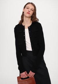 Cardigan nero lavorato a maglia con chiusura a bottoni, dotato di due tasche frontali e polsini a coste, abbinato a una parte superiore bianca e con in mano una borsa marrone.