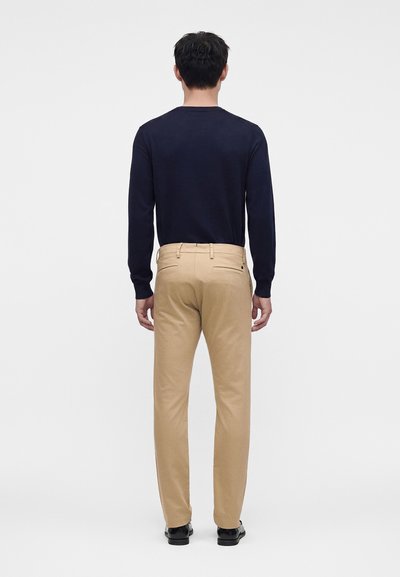 Mand, der står med ryggen til iført en marineblå langærmet skjorte, beige bukser og sorte sko mod en ensfarvet hvid baggrund.