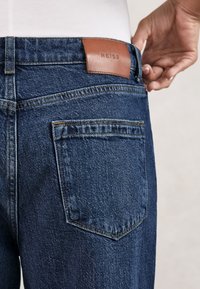 Close-up af blå denimjeans baglomme med brun læder "REISS" mærkat og en hånd, der justerer taljen.