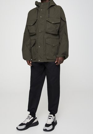 Homme portant une veste à capuche verte olive avec plusieurs poches avant, un pantalon noir et des baskets noir et blanc, debout devant un fond uni.