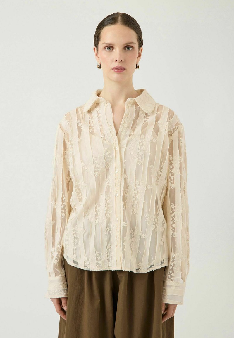 Žena s kosom počešljanom prema nazad, odjevena u kremastu prozirnu bluzu s cvjetnim vezom i široke smeđe hlače, okrenuta prema naprijed na jednobojnoj pozadini.