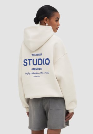 Frau mit dunklen Haaren zu einem Dutt gebunden, trägt einen weißen Hoodie mit blauem "STUDIO GARMENTS"-Schriftzug auf dem Rücken und graue Shorts, seitlich stehend.
