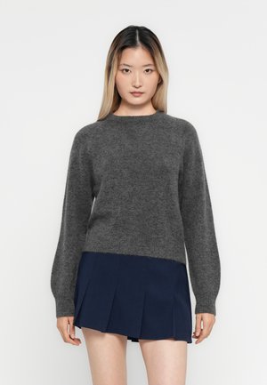 Junge Frau mit langen Haaren, die einen dunkelgrauen Strickpullover und einen marineblauen, plissierten Rock trägt, steht vor einem schlichten weißen Hintergrund.