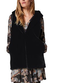 Femme portant un gilet à capuche noir sans manches par-dessus une robe à manches longues à motif cachemire, les mains dans les poches du gilet.