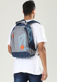 Zaino in tessuto grigio con motivo geometrico blu, zip arancio, tasca laterale in rete e doppie spalline. Adatto per l'uso quotidiano.