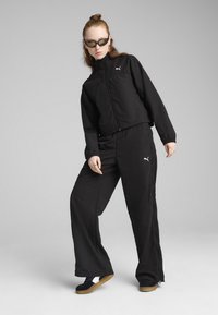 Črn trenirkar z zadrgo in širokimi hlačami. Značilni so elastični manšeti in bel logotip Puma. Uporabljen z črnimi čevlji.