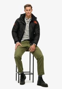 Mężczyzna siedzący na stołku, ubrany w czarną kurtkę z kapturem z pomarańczowym logo, szary podkoszulek typu henley, oliwkowe spodnie i czarne botki na sznurówki.