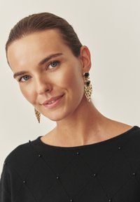 Goldfarbene Ohrringe mit einem Blattdesign, die mit schwarzen Akzentsteinen verziert sind. Das Model trägt einen schwarzen Pullover mit einem strukturierten, diamantförmigen Muster.