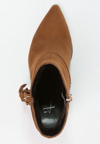 Hnědé semišové kotníkové boty s špičatou špičkou, bočním zipem a dekorativními třásněmi, zobrazené z pohledu shora na bílém pozadí.