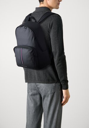 Zaino nero con una texture liscia, dotato di una tasca frontale con un accento a strisce colorate e spallacci regolabili.