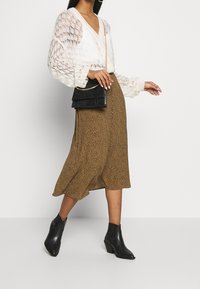 Krémový pletený cardigan s výstřihem do V a zapínáním na knoflíky, kombinovaný s midi sukní hnědé barvy s puntíky a černými kotníkovými botami. Černá kabelka jako doplněk.