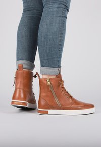 Botas de tobillo de cuero color marrón claro con un acabado texturizado, detalle de cremallera dorada y suela blanca. Jeans de mezclilla enrollados visibles por encima de las botas.