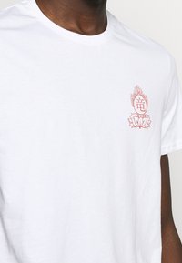 Bílý bavlněný tričko s červeným květinovým vzorem a symbolem na levé části hrudi. Klasický kulatý výstřih a krátké rukávy.