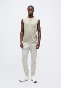 Άνδρας που στέκεται, φορώντας μπεζ αμάνικο τοπ, ανοιχτό γκρι παντελόνι τζόγκινγκ και λευκά αθλητικά παπούτσια μπροστά σε απλό ανοιχτόχρωμο φόντο.